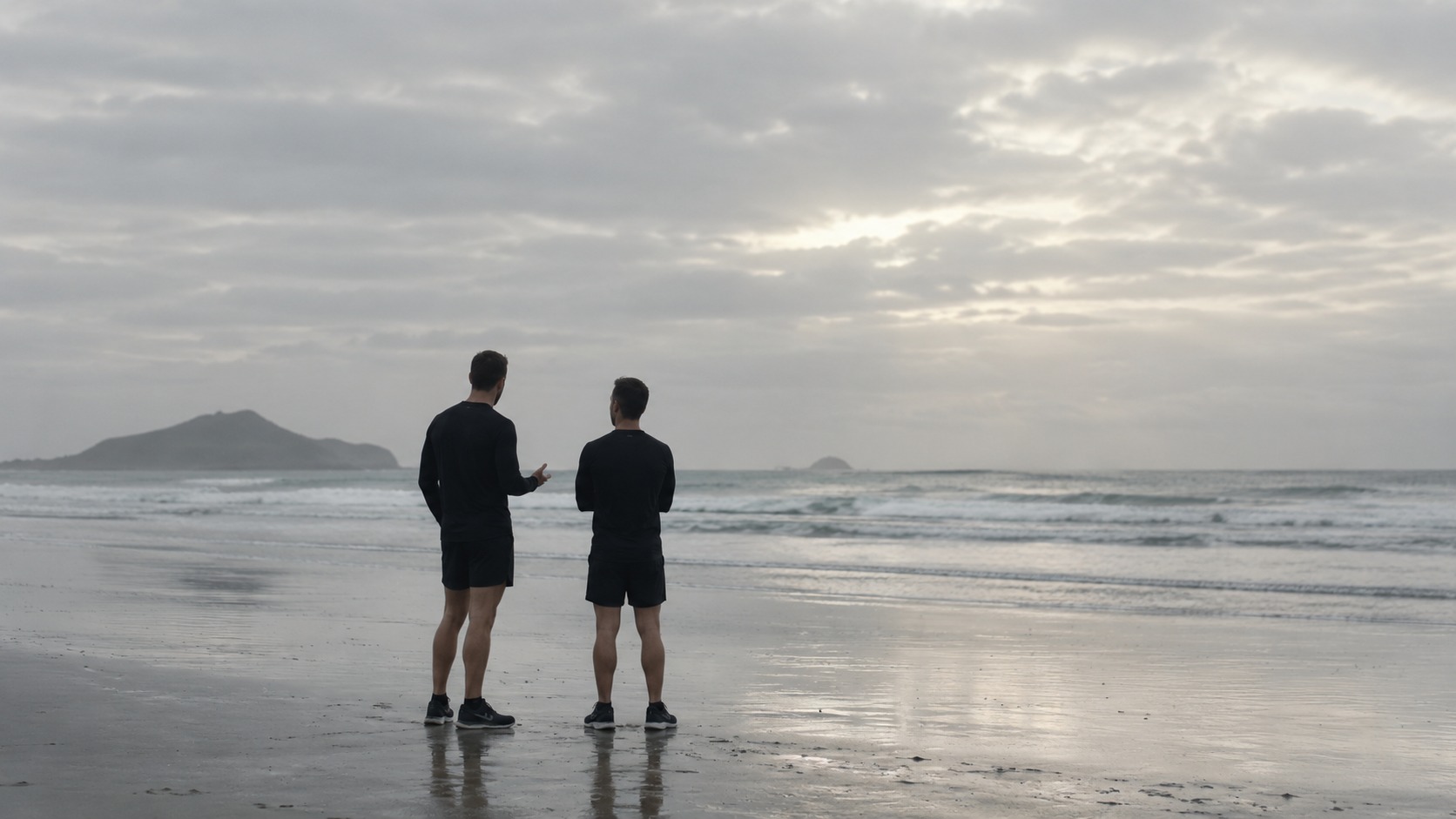 Two men on the sand at dawn, backs to camera, one gesturing mid conversation.